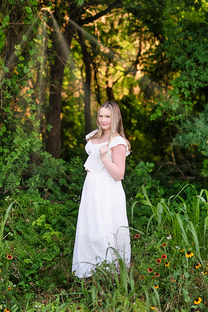 Senior girl in a white dress posing among lush trees for her senior portraits in Fort Worth